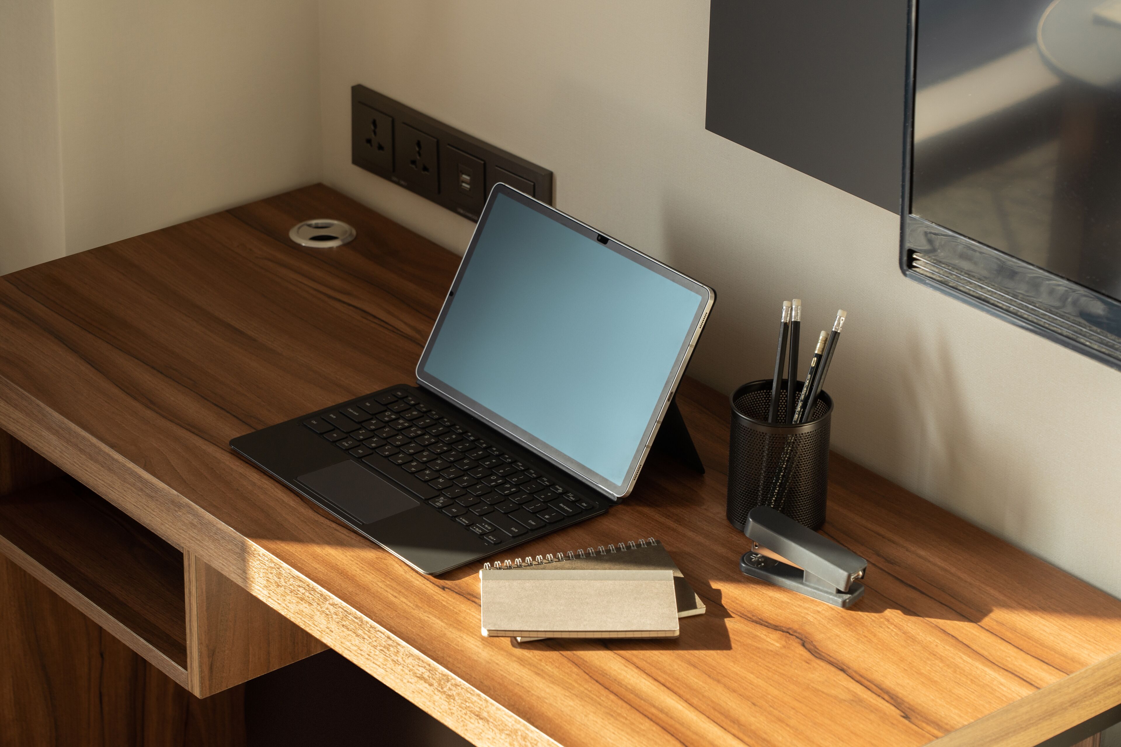 A wooden desk with a laptop, a notepad, a stapler, and a pen holder.
