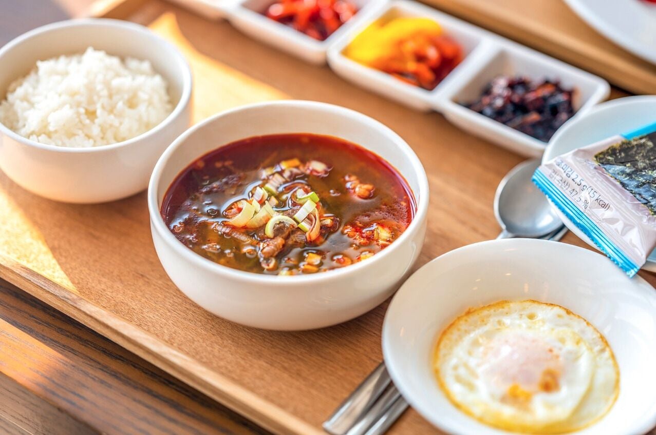 Un plateau repas comprenant du riz, un bol de soupe, un œuf frit et divers accompagnements.