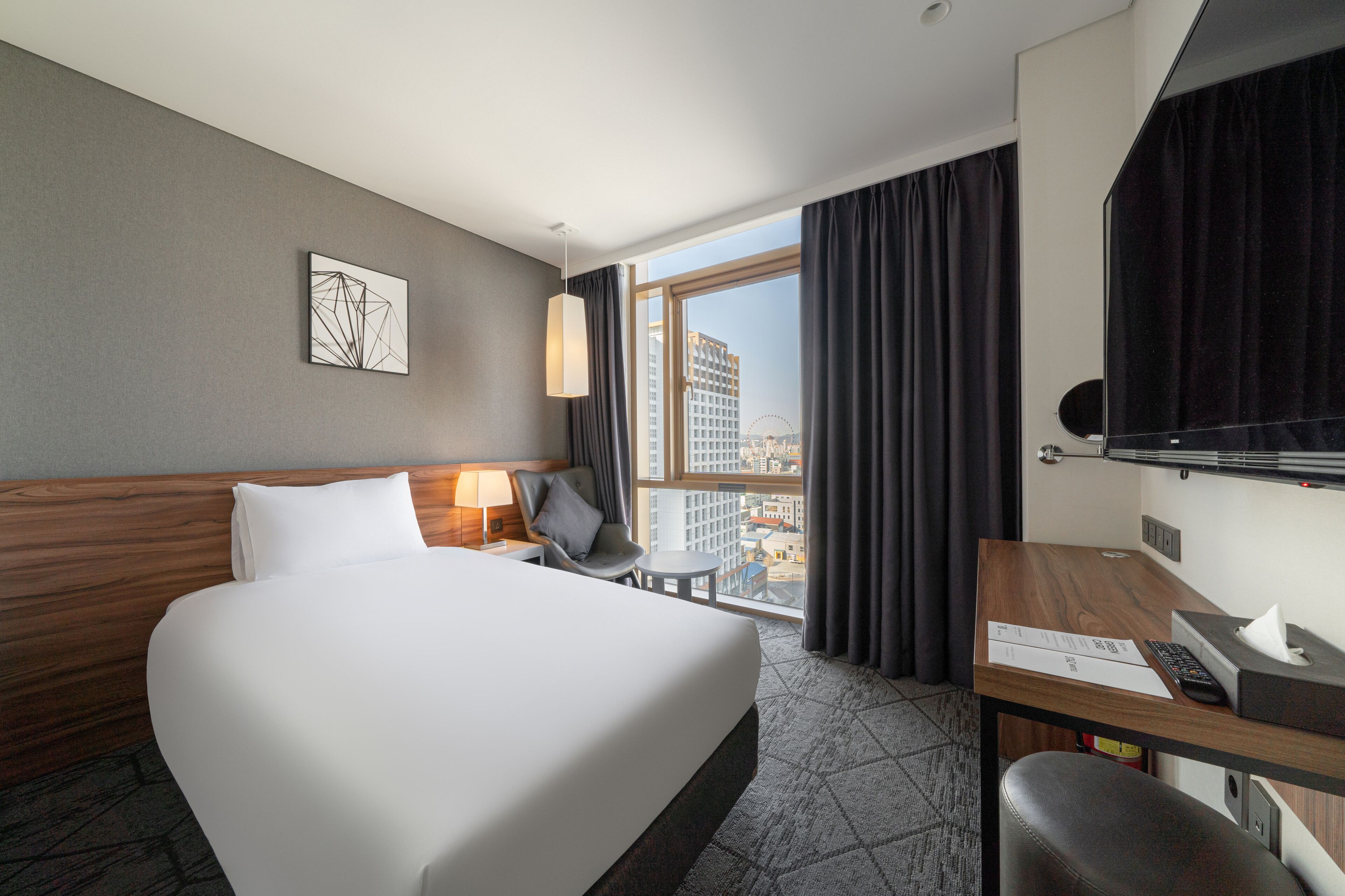 A hotel room with a large bed, a desk with a computer, a chair, a lamp, and a window with curtains.