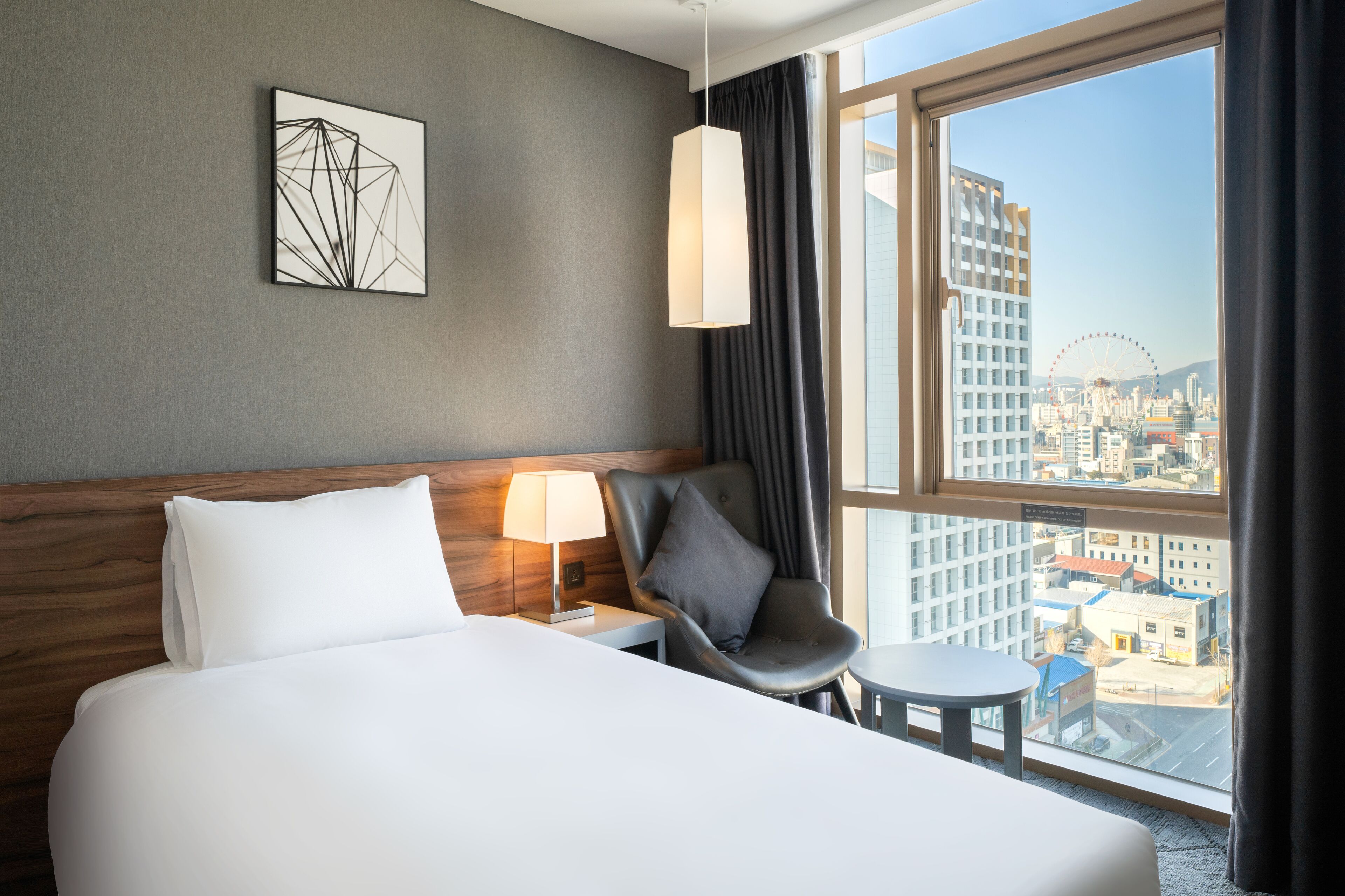 Hotellihuone, jossa on sänky, tuoli, pieni pöytä, lamppu ja suuret ikkunat, joista avautuu näkymä kaupunkiin.
