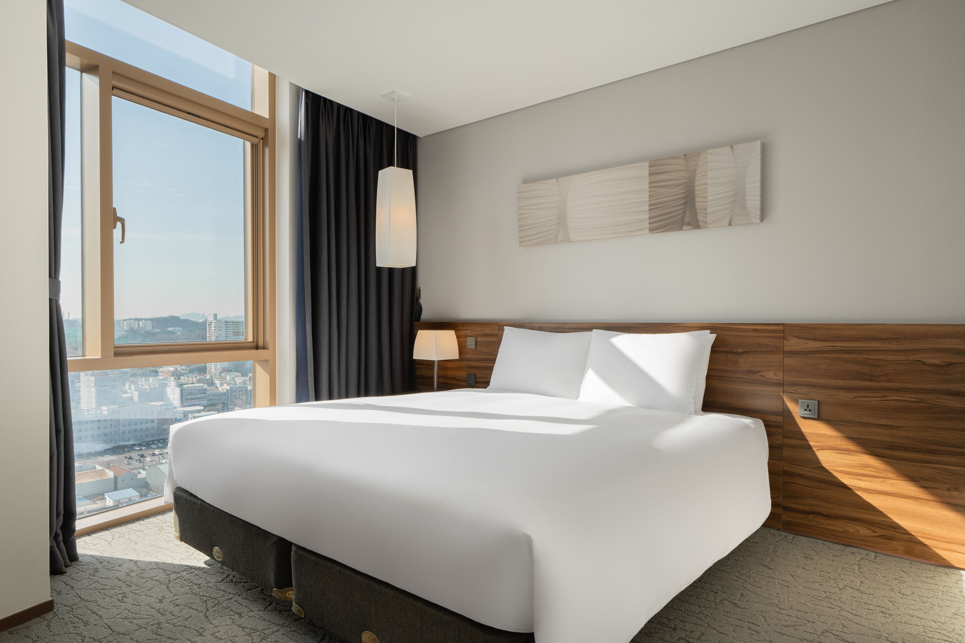A hotel room with a large bed, a city view, a modern lamp, and a wooden headboard.