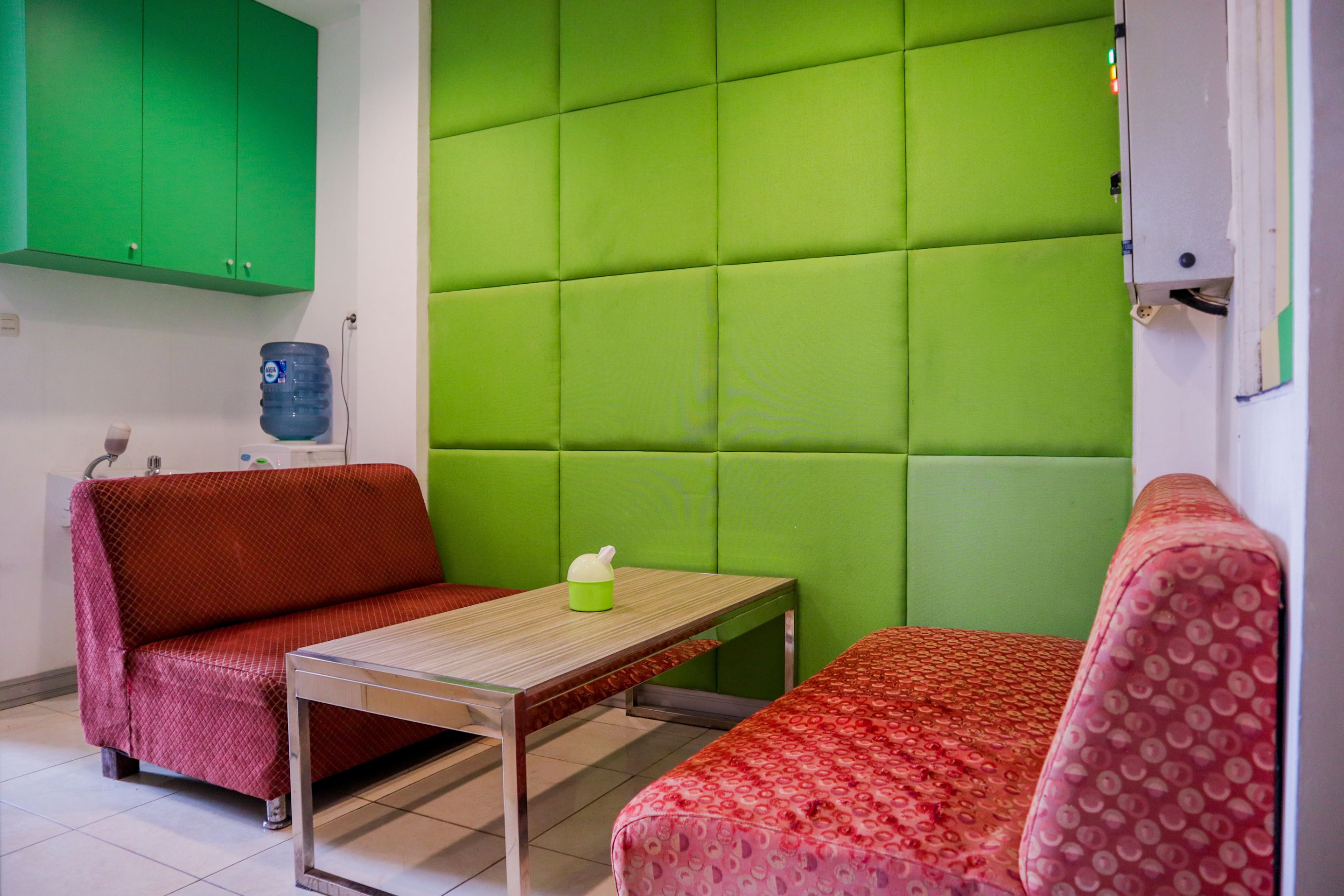 A small room with a green padded wall, a red sofa, a wooden table, and a red patterned sofa.