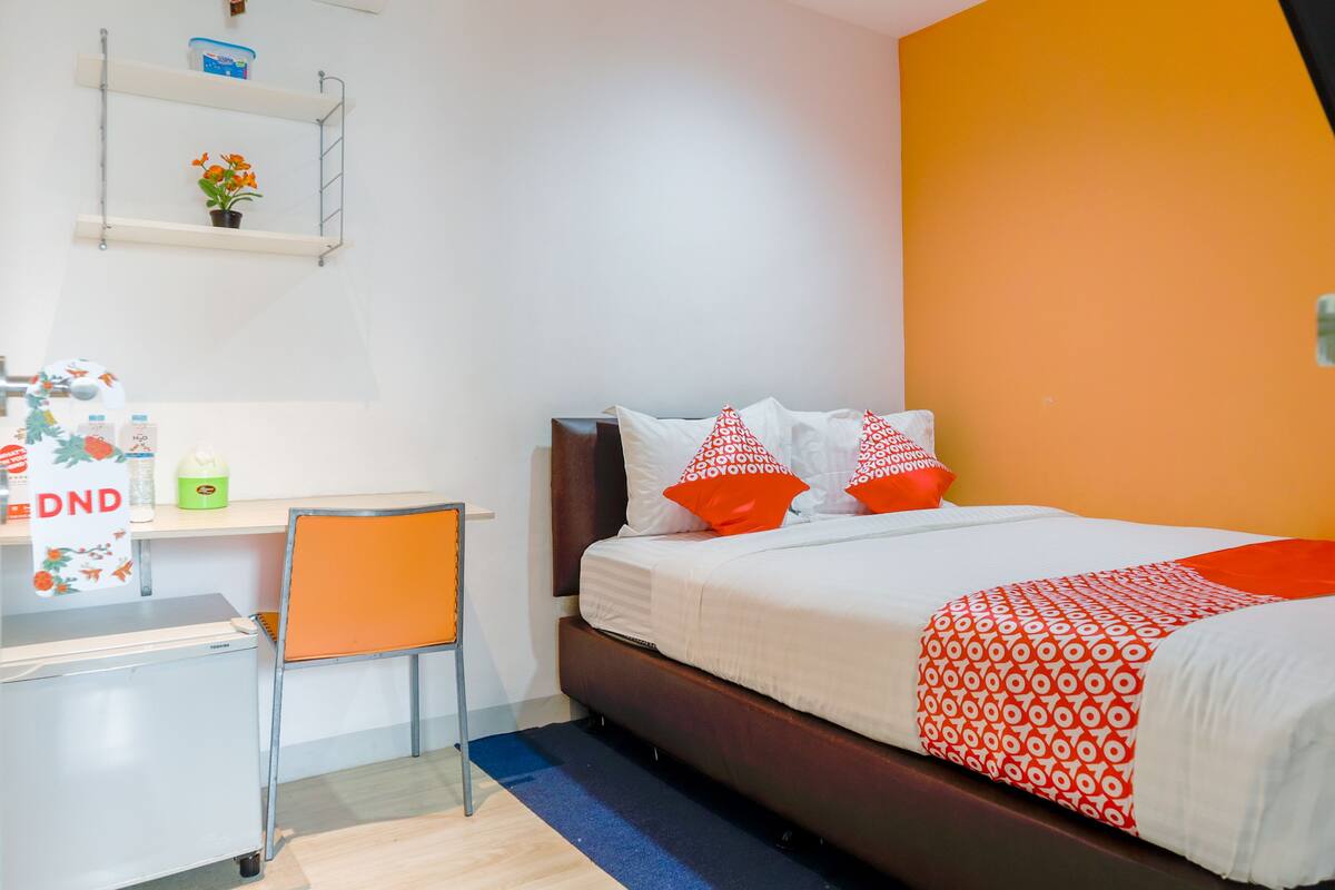 Ein Hotelzimmer mit einem Bett, einem Schreibtisch mit Stuhl, einem Regal mit Pflanze und einem blauen Teppich.
