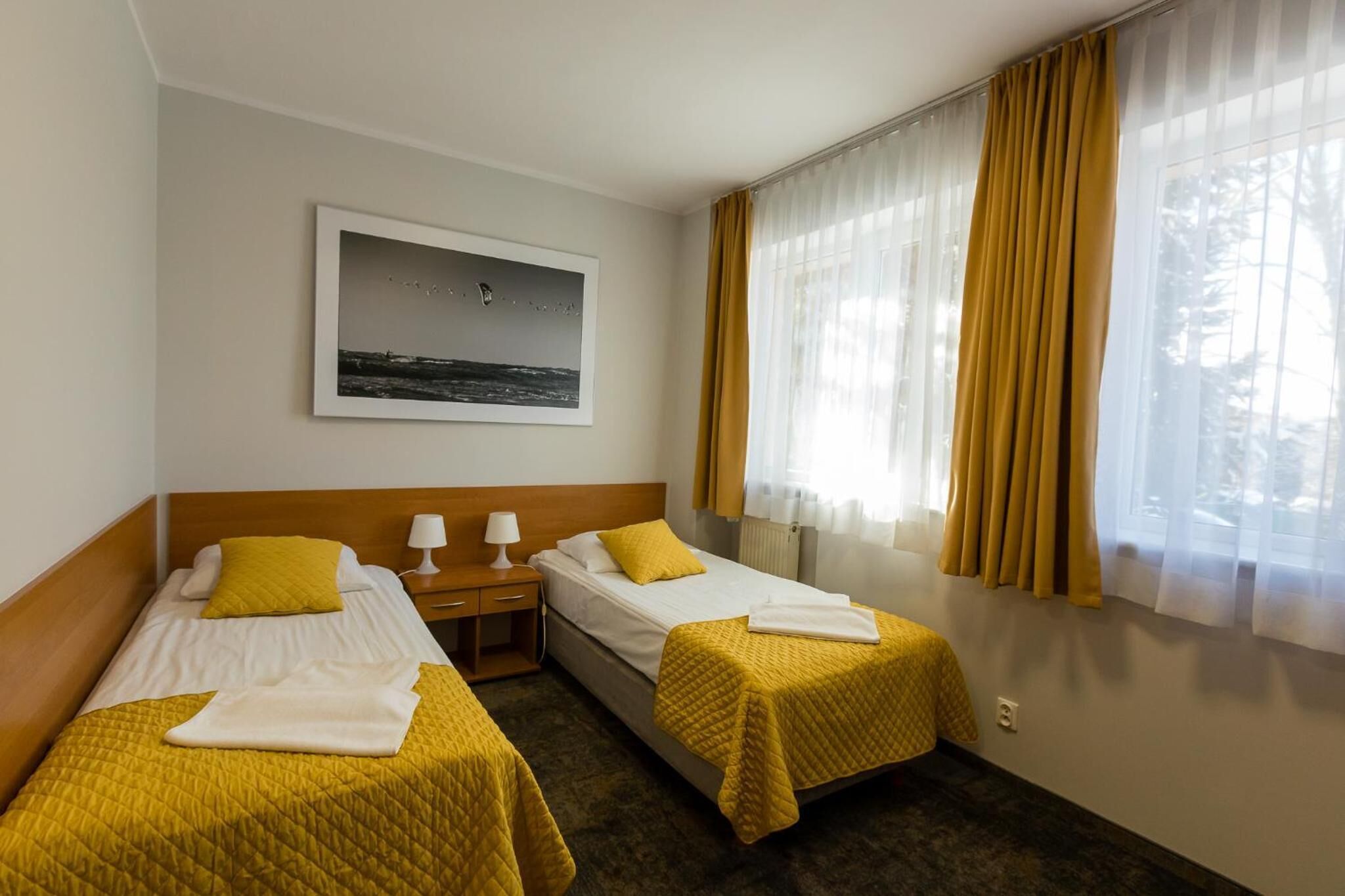Ein Hotelzimmer mit zwei Betten, einem Nachttisch, einer Lampe, einem gerahmten Bild an der Wand und einem Fenster mit Vorhängen.