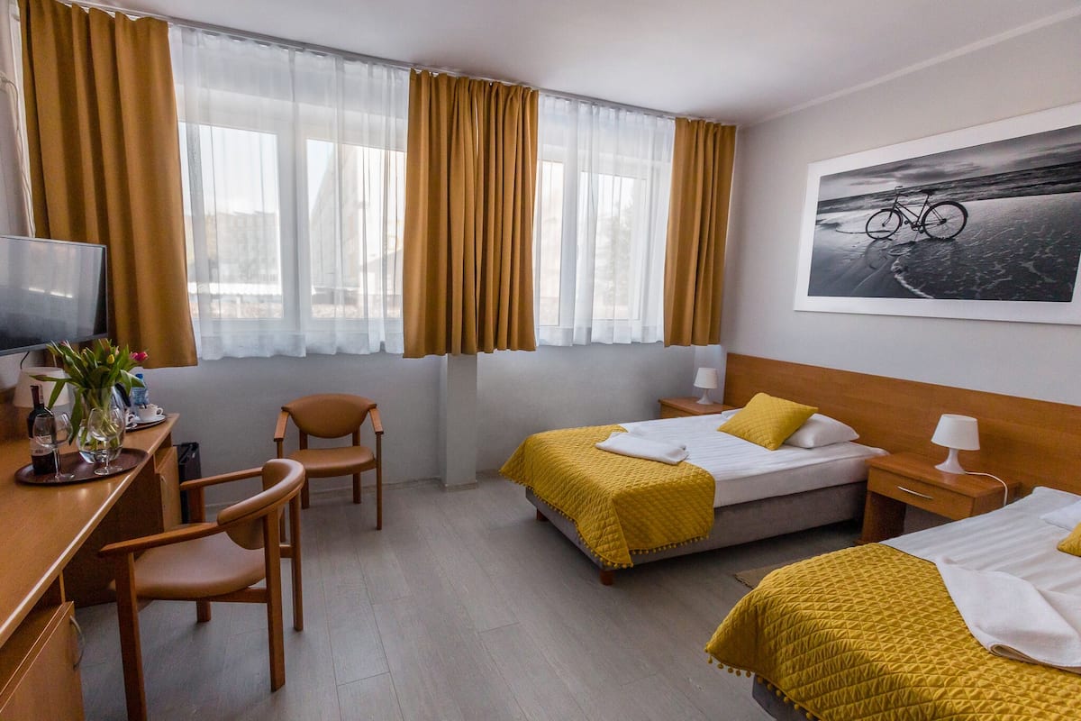 A hotel room with two beds, a desk with a chair, a television, and a painting of a bicycle on the wall.