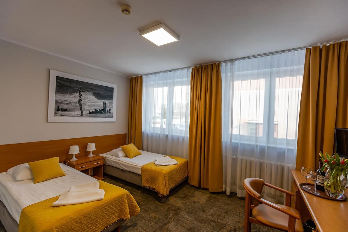 A hotel room with two beds, a desk with a chair, a painting on the wall, and a window with curtains.