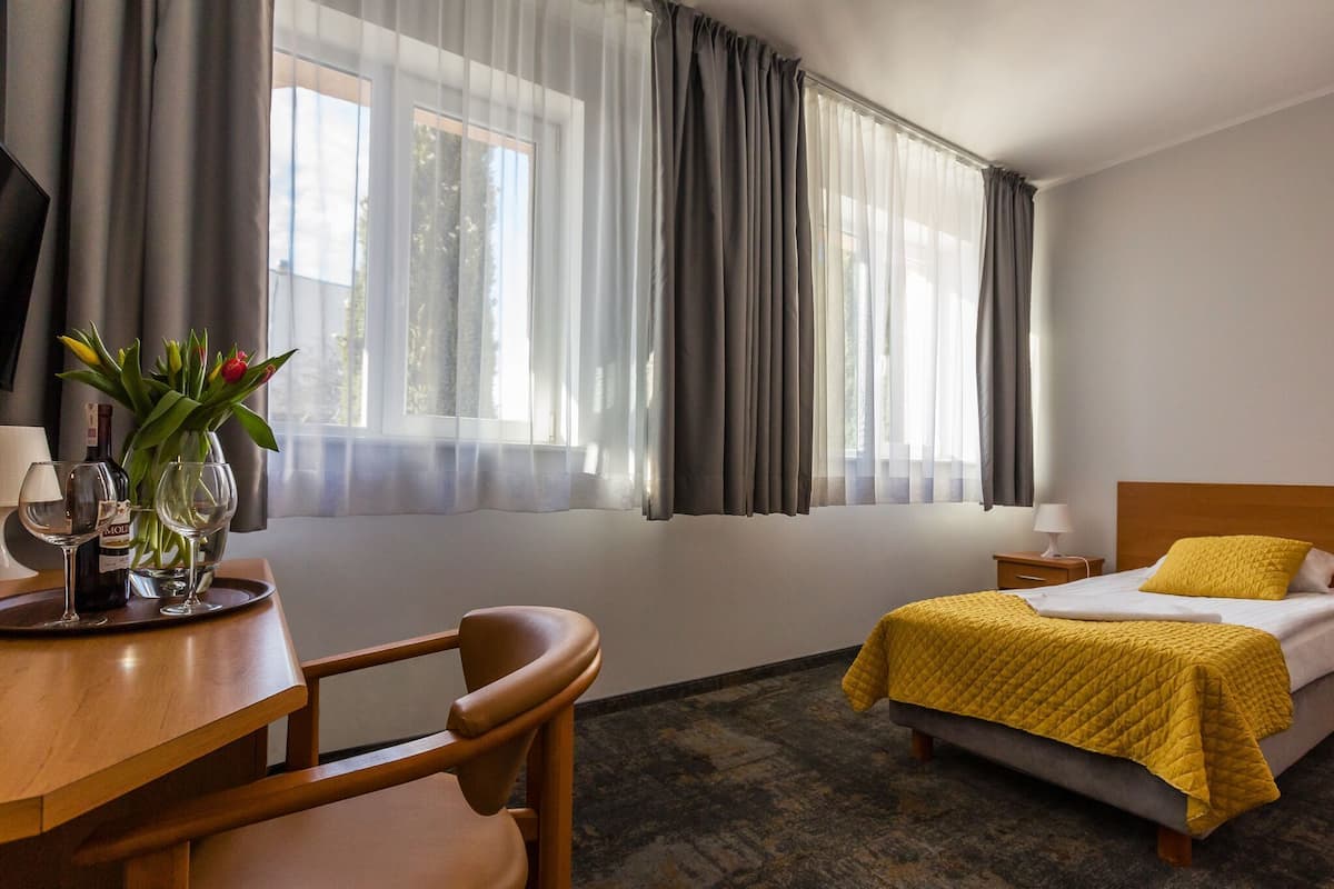 A hotel room with a bed, a desk with a chair, a vase with flowers, and a window with curtains.