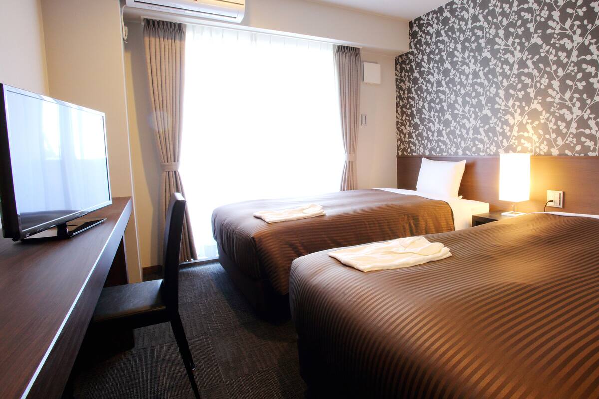 A hotel room with two beds, a desk with a flat-screen TV, an air conditioner, and a floral-patterned wall.