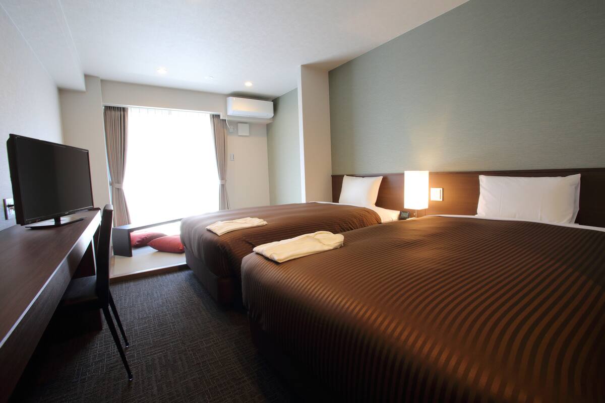 A hotel room with two beds, a desk, a television, and a window with curtains.
