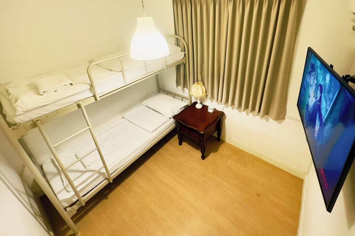 A bunk bed room with a bedside table, a lamp, a TV, and a window with curtains.
