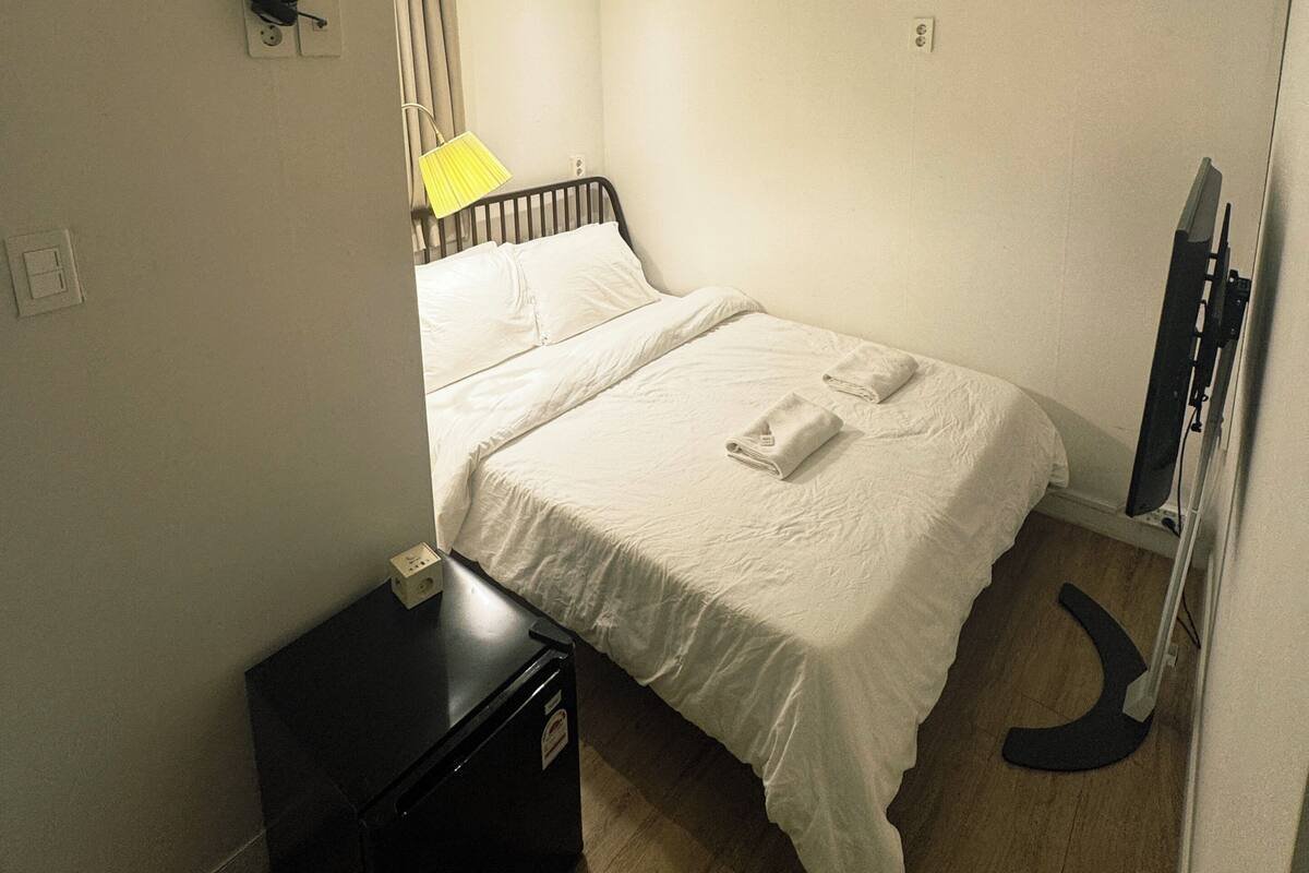 A hotel room with a bed, a desk, a chair, a flat-screen TV, and a ceiling-mounted air conditioner.