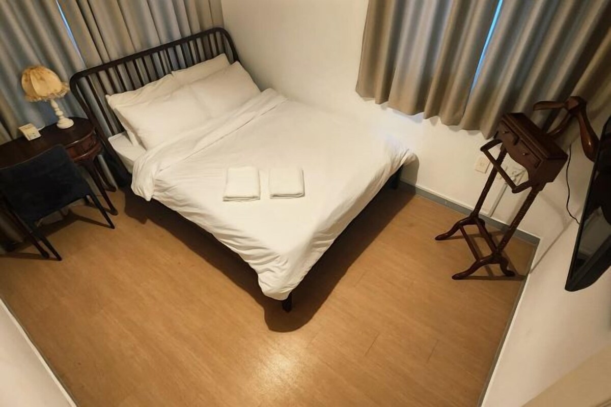Een net opgemaakt bed met witte lakens, een nachtkastje met een lamp en een kleine houten stoel.