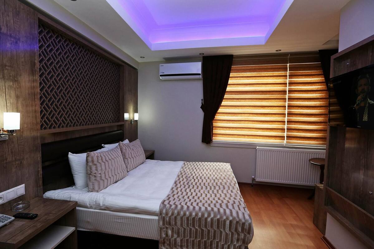 Habitación de hotel con cama, mesita de noche, lámpara, aire acondicionado, ventana con persianas y un foco en el techo.