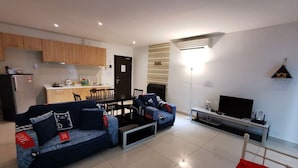 A modern living room with a sofa, a TV, and a dining area with wooden cabinets and a kitchen in the background. - Homestay in Johor - Bear House (Johor Bahru)