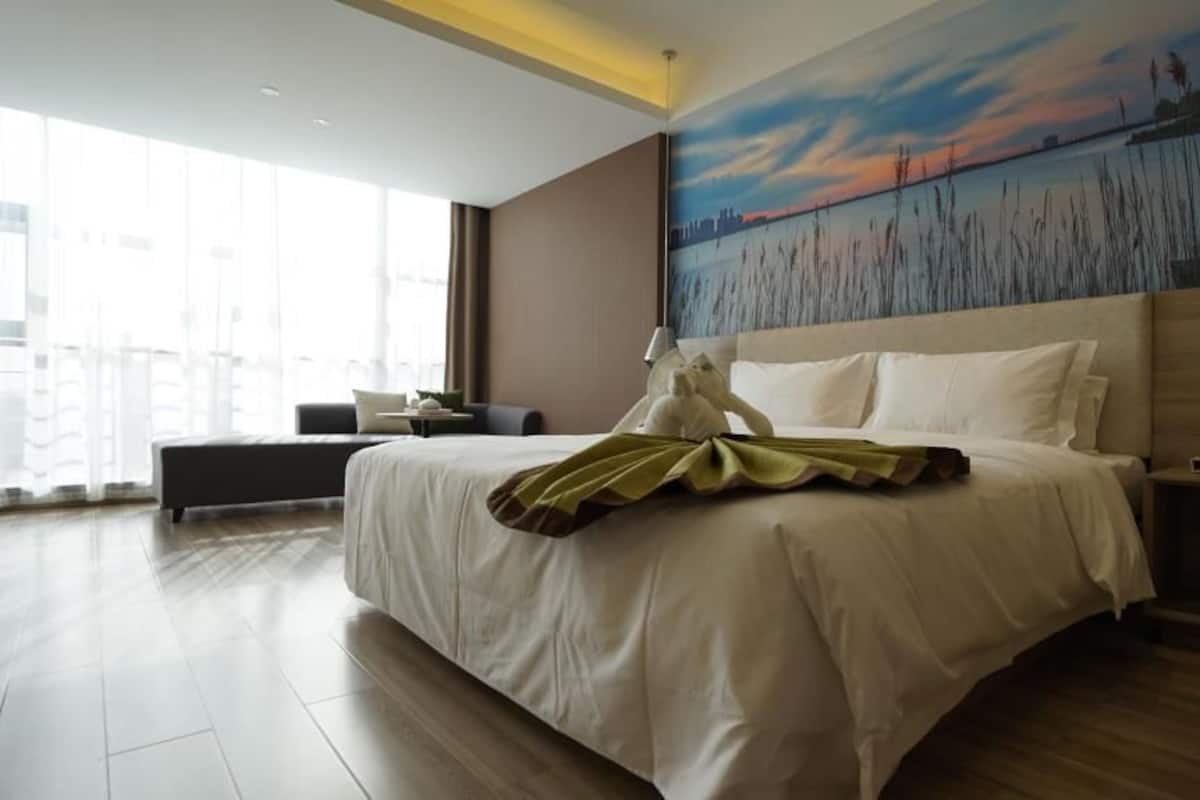 A modern hotel room with a large bed, a nightstand, a desk, and a view of a sunset over water.