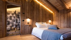 A wooden cabin bedroom with a bed, bedside lamps, and a wooden door leading to a bathroom with a hexagonal tile backsplash. - Lavarets Chambres d'Hotes (Ayas)