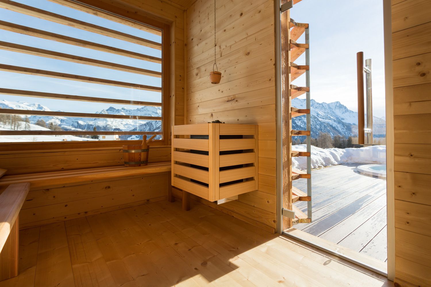 A wooden cabin with a wooden bench, a hanging basket, and a view of snow-covered mountains.