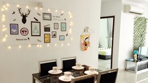 Uma sala de jantar com uma mesa posta para quatro pessoas, uma parede decorada com quadros e luzes de cordão, e uma cama visível ao fundo.