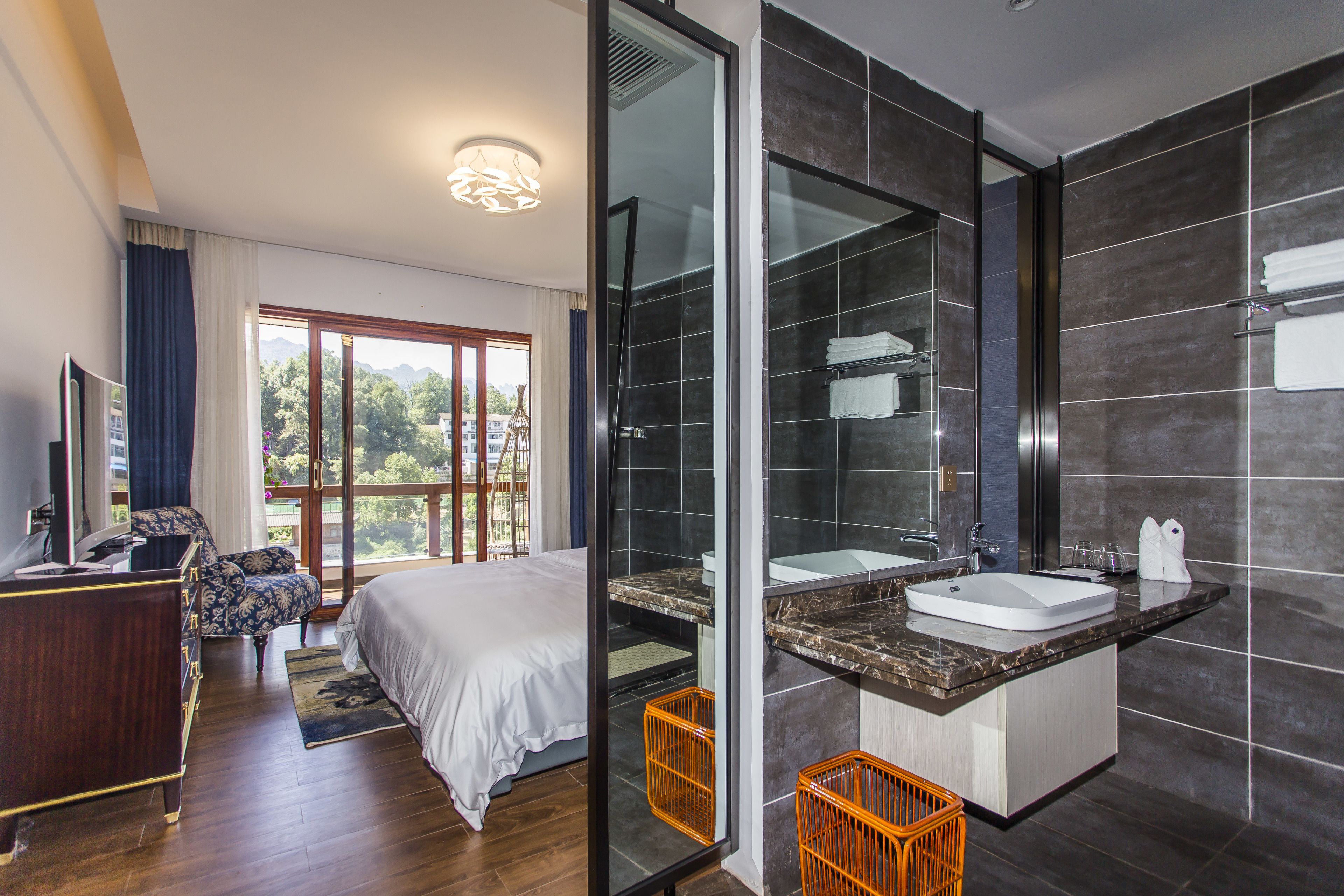 Habitación de hotel moderna con una cama grande, un escritorio y un baño con un espejo grande, lavamanos y azulejos oscuros.