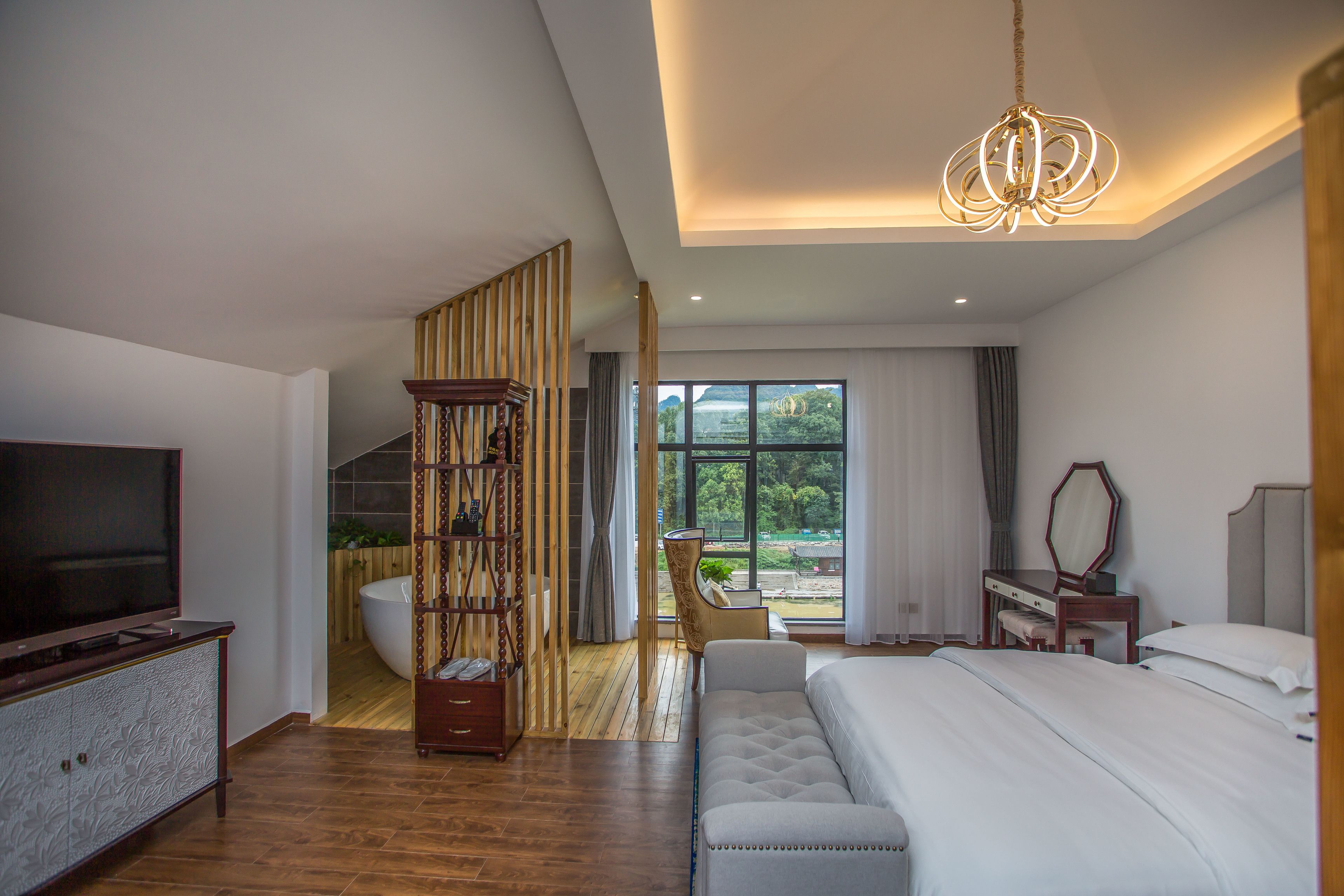 A modern hotel room with a large bed, a TV, a wooden bookshelf, a desk, and a view of the outdoors.