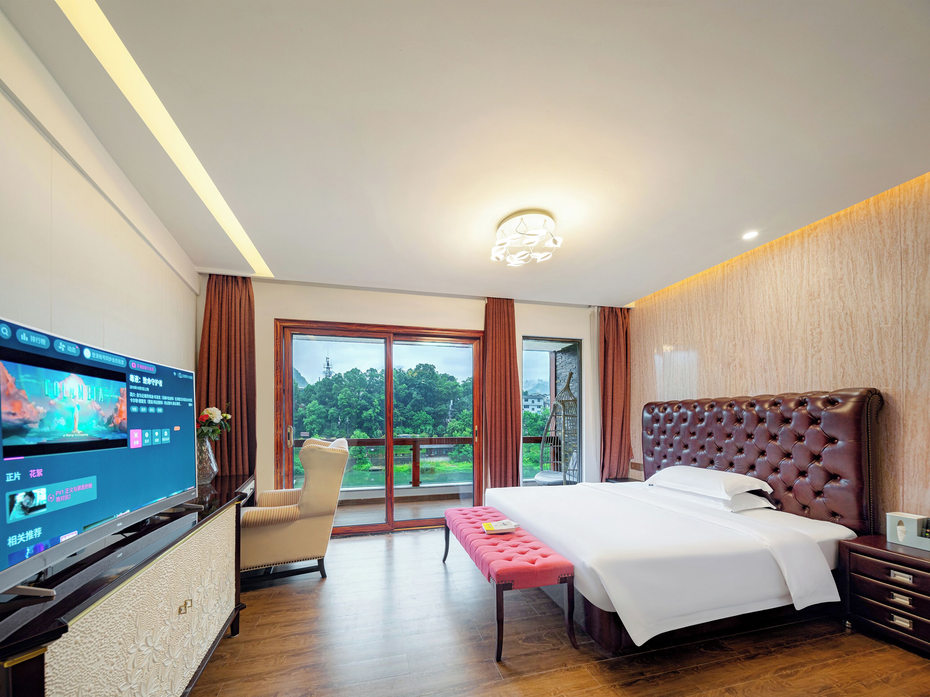 A hotel room with a large TV, a bed, a bench, a chair, and a view of greenery outside.