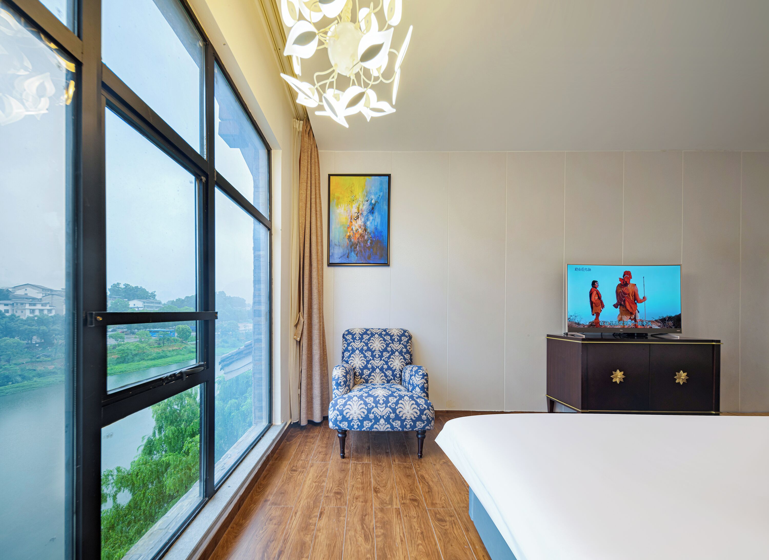 Ein Hotelzimmer mit großem Fenster, einem blau gemusterten Sessel, einem Fernseher und einem Bett.