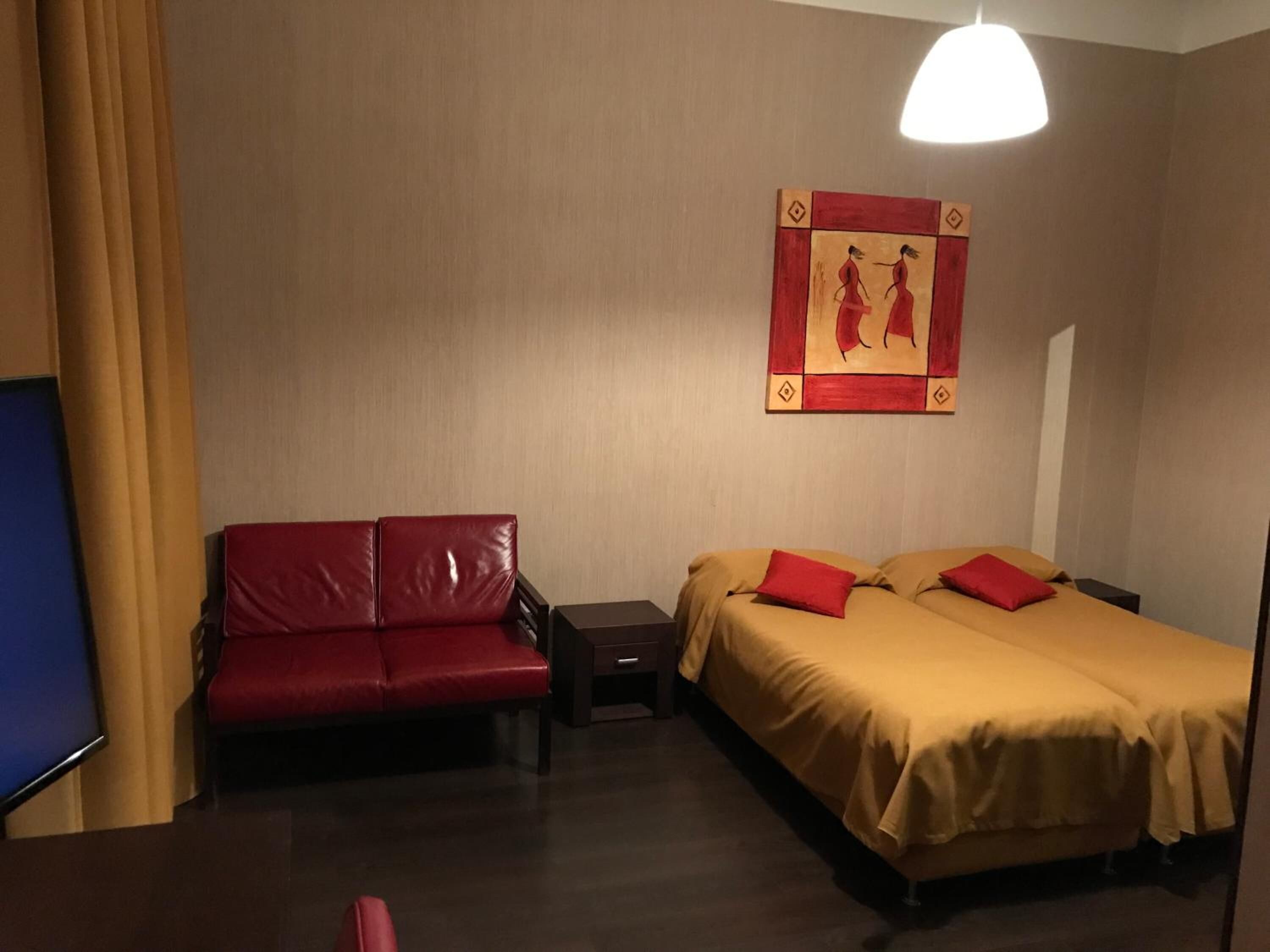 A hotel room with a red leather sofa, a double bed with red pillows, a nightstand, and a wall-mounted artwork.