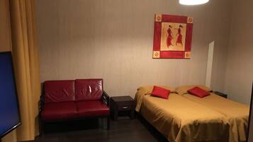 A hotel room with a red leather sofa, a double bed with red pillows, a nightstand, and a wall-mounted artwork.