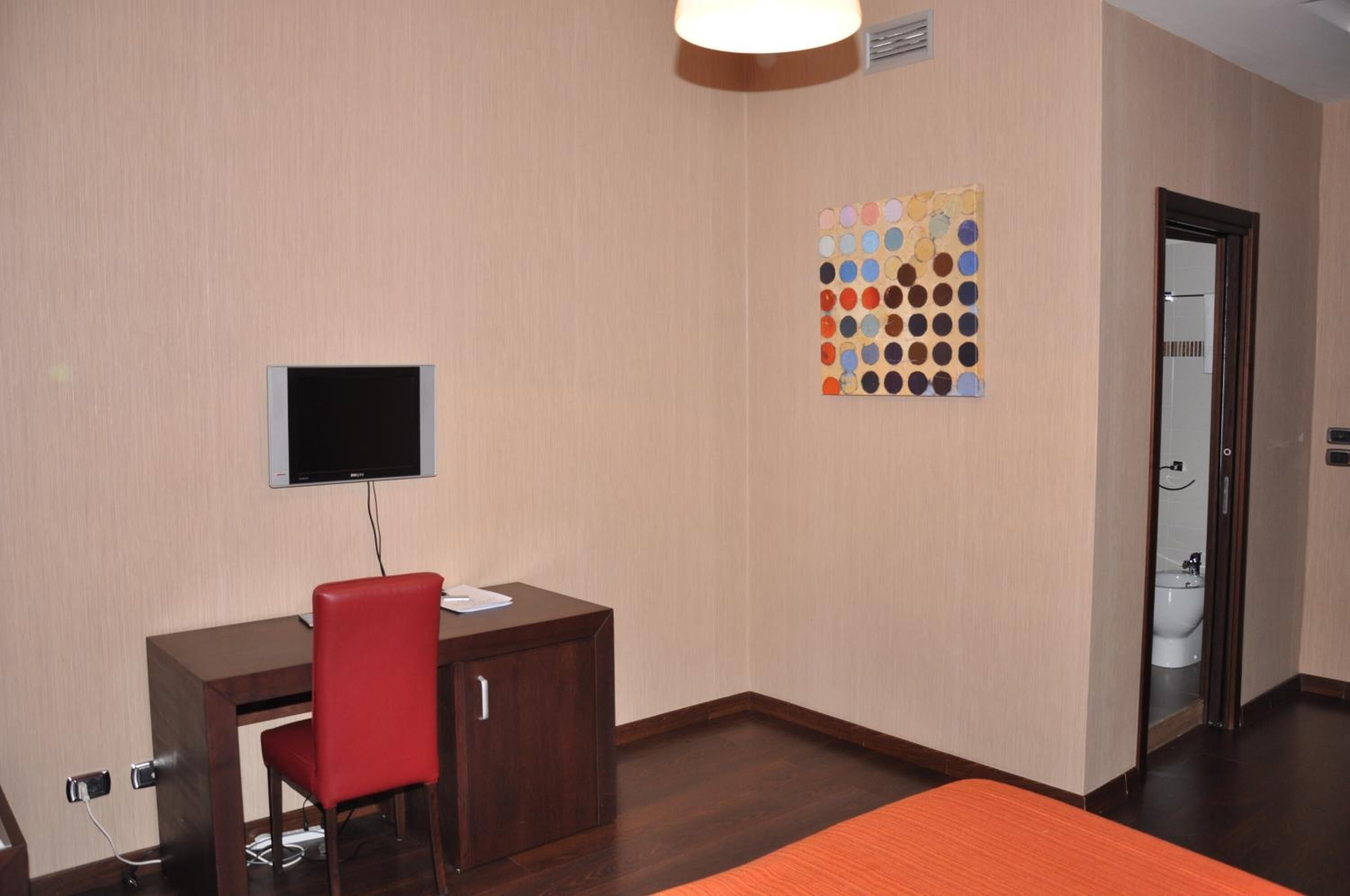 Habitación de hotel con escritorio, una silla roja, televisor montado en la pared y una obra de arte colorida.