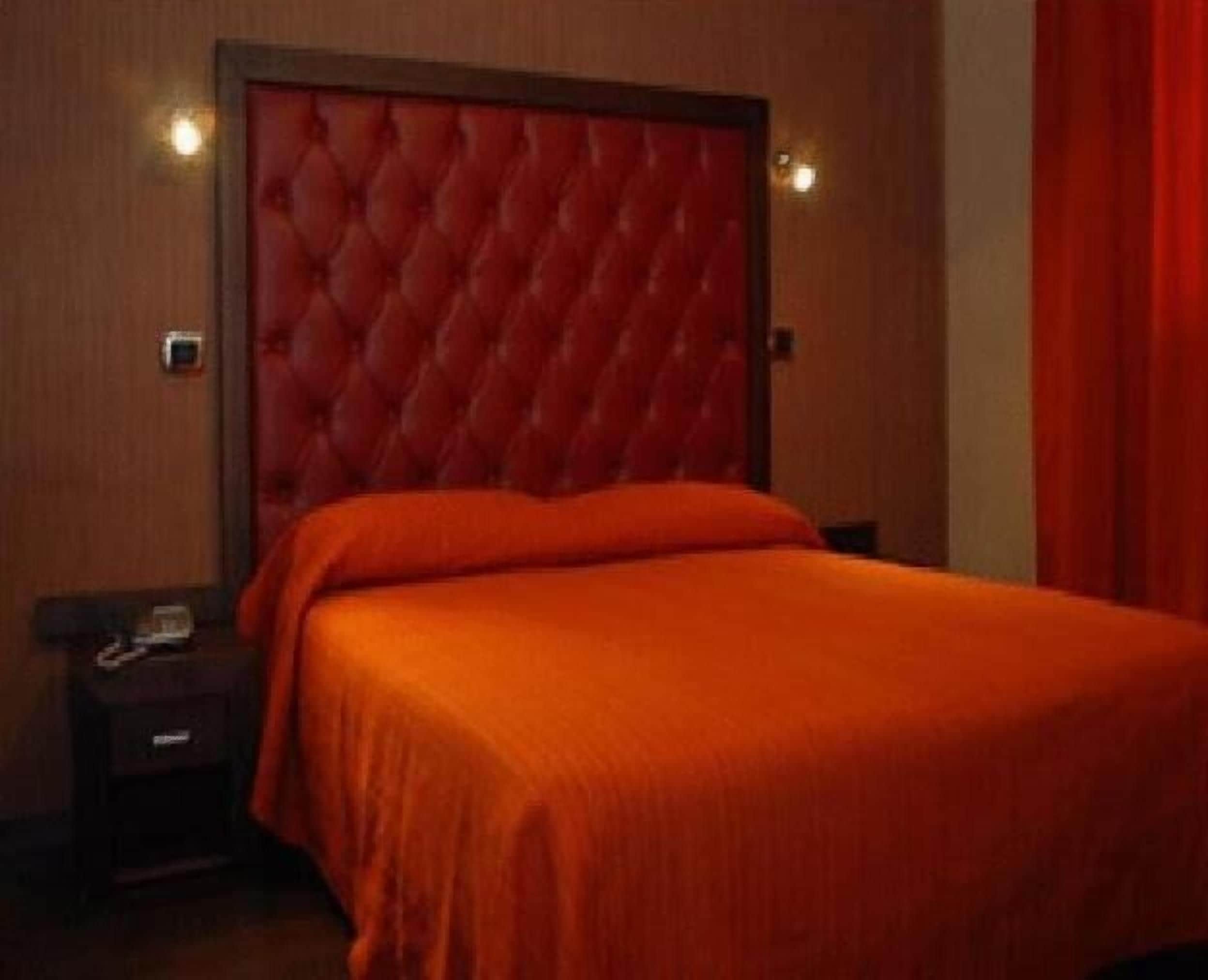 A hotel room with a tufted headboard, a single bed with an orange bedspread, a nightstand with a phone, and a red curtain.