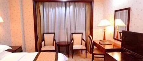 A hotel room with a bed, two chairs, a desk with a lamp, a dresser, and a large window with curtains.