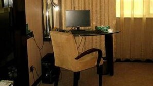 Quarto de hotel com uma cama de solteiro, uma mesa com computador, uma cadeira e uma televisão.
