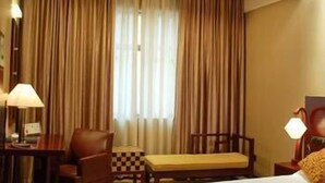 Quarto de hotel com cama, escrivaninha, cadeira, abajur e janela com cortinas.