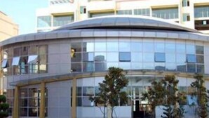 Um edifício moderno com fachada curva, várias janelas e um jardim frontal ajardinado com árvores e arbustos.