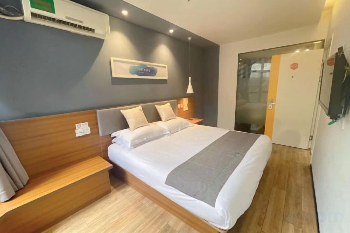 Une chambre d’hôtel comprenant un lit, des tables de chevet, un climatiseur fixé au mur, un tableau encadré et une porte avec un panneau en verre.