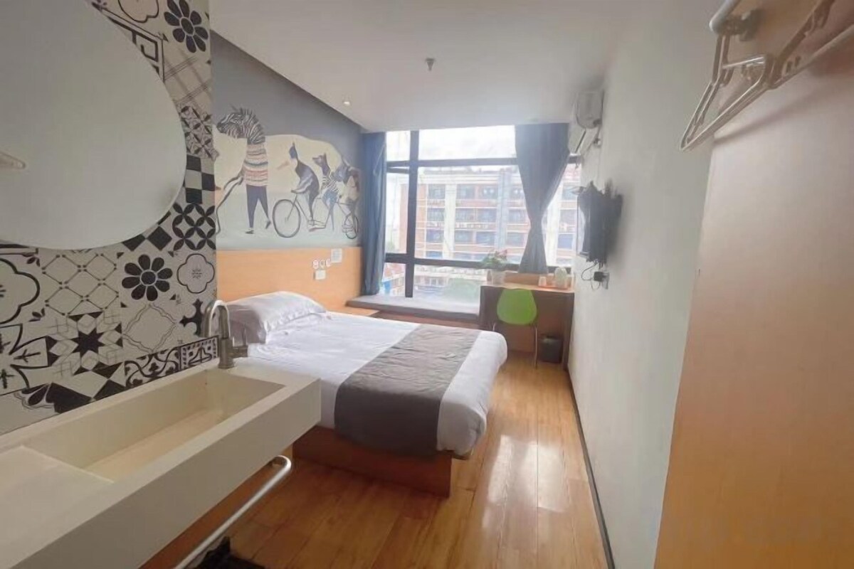 A hotel room with a bed, a large window, a desk, and a wall-mounted TV.