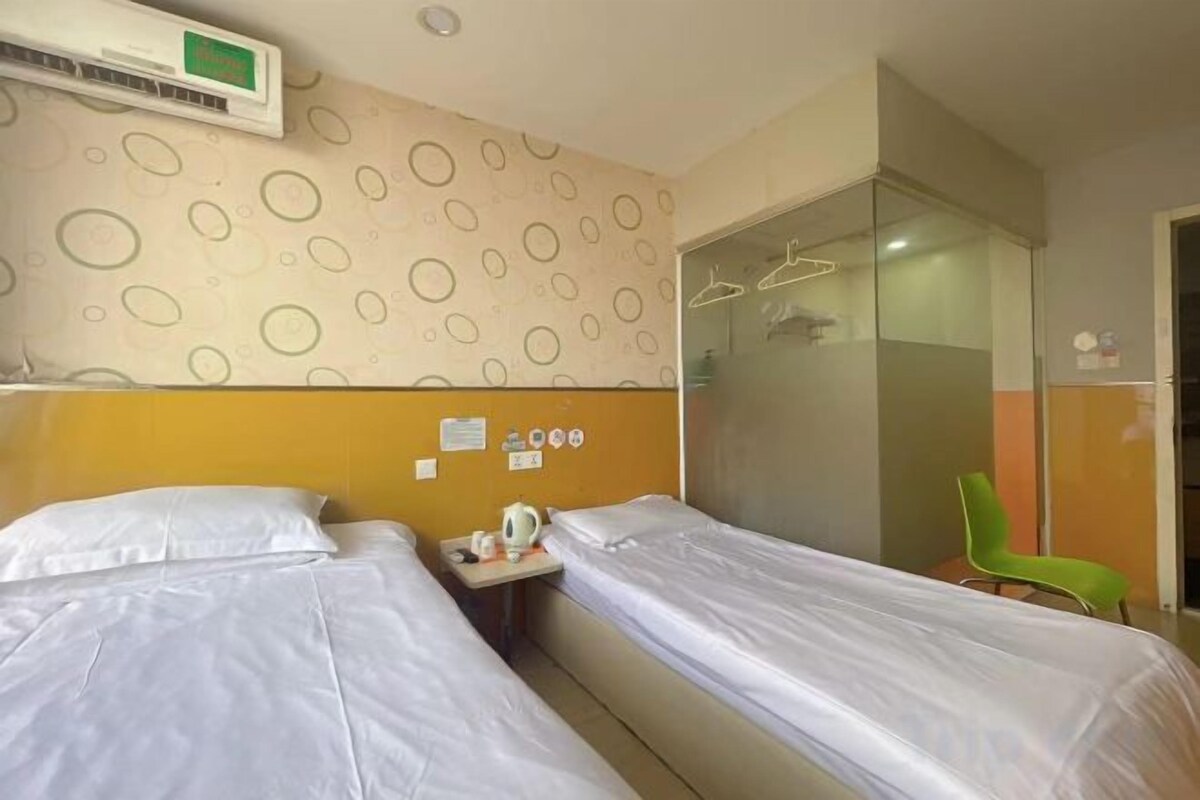 A hotel room with two beds, a small table with a kettle, a mirror, and a green chair.