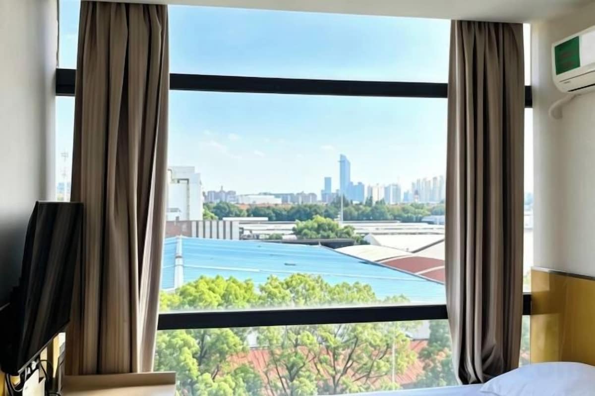 A hotel room with a large window, curtains, a bed, and a TV.