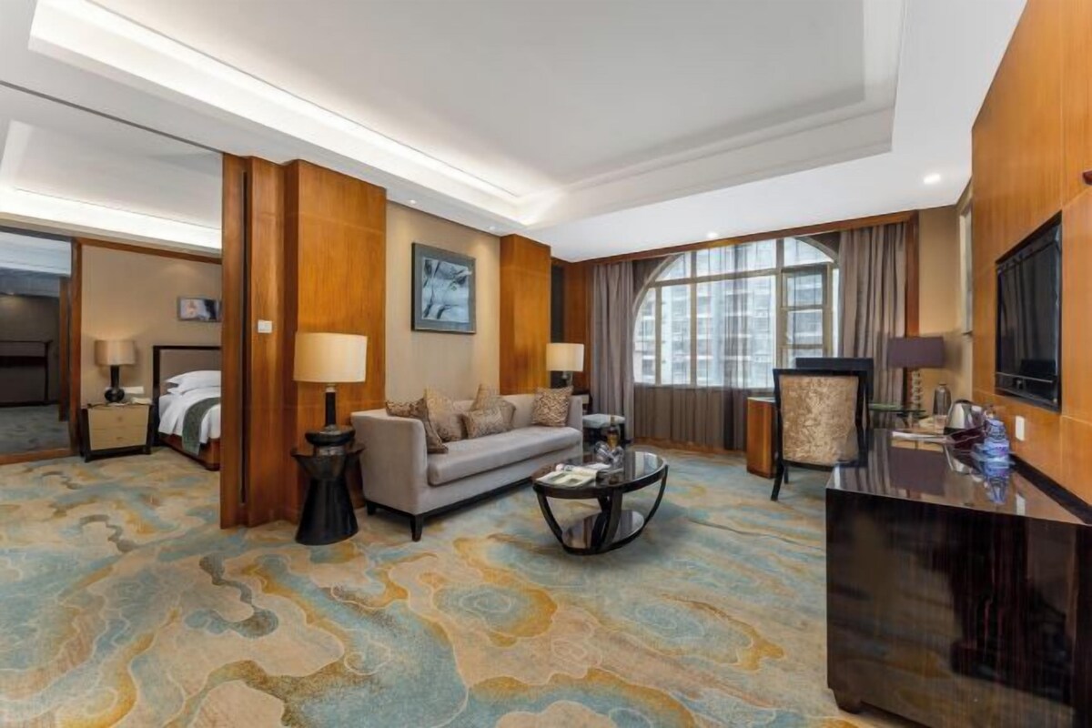 A hotel room with a sofa, two armchairs, a coffee table, a TV, and a large window with curtains.