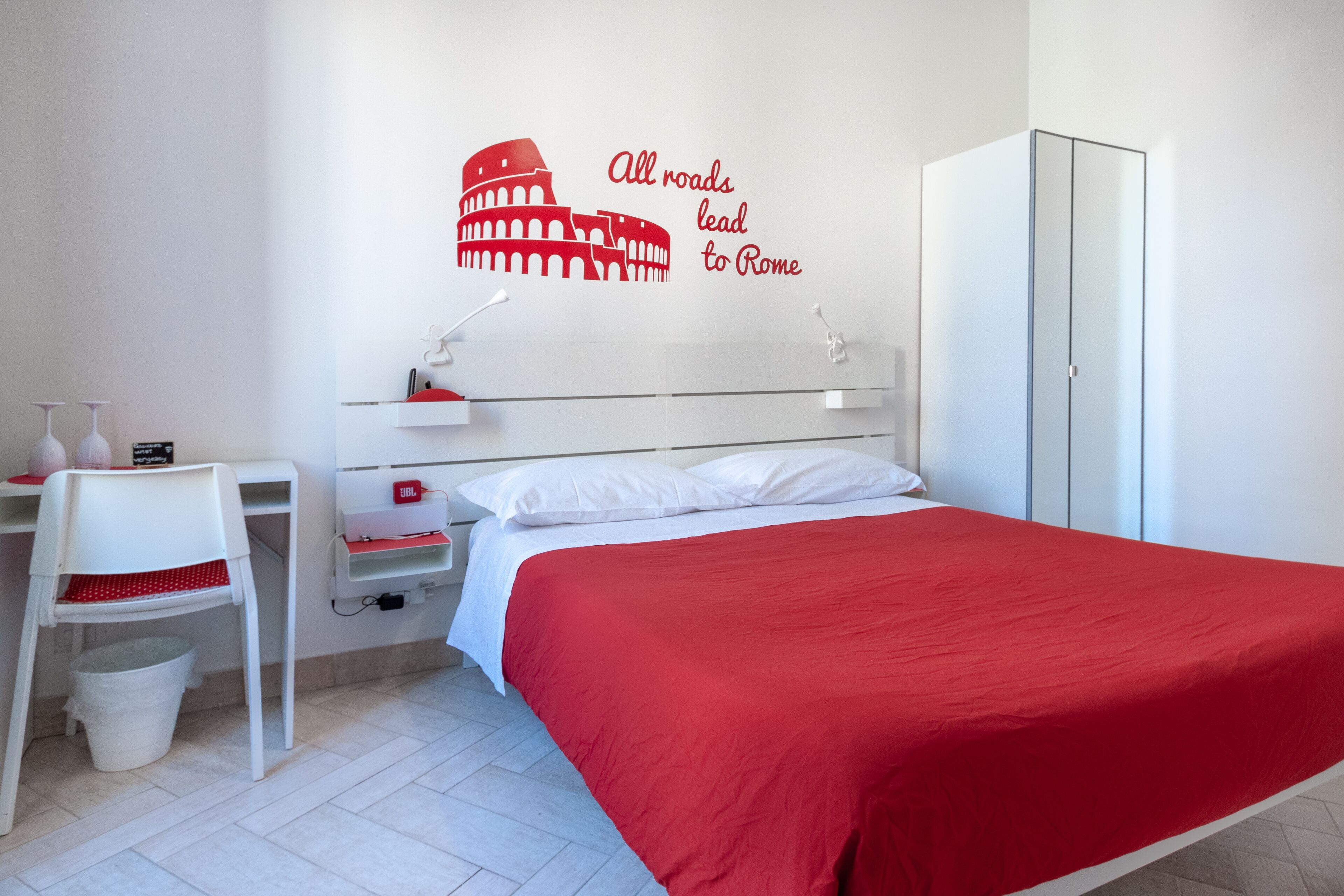 Une chambre à coucher comprenant un lit, un bureau, une chaise, une armoire et un autocollant mural représentant le Colisée, avec le texte : « Toutes les routes mènent à Rome ».