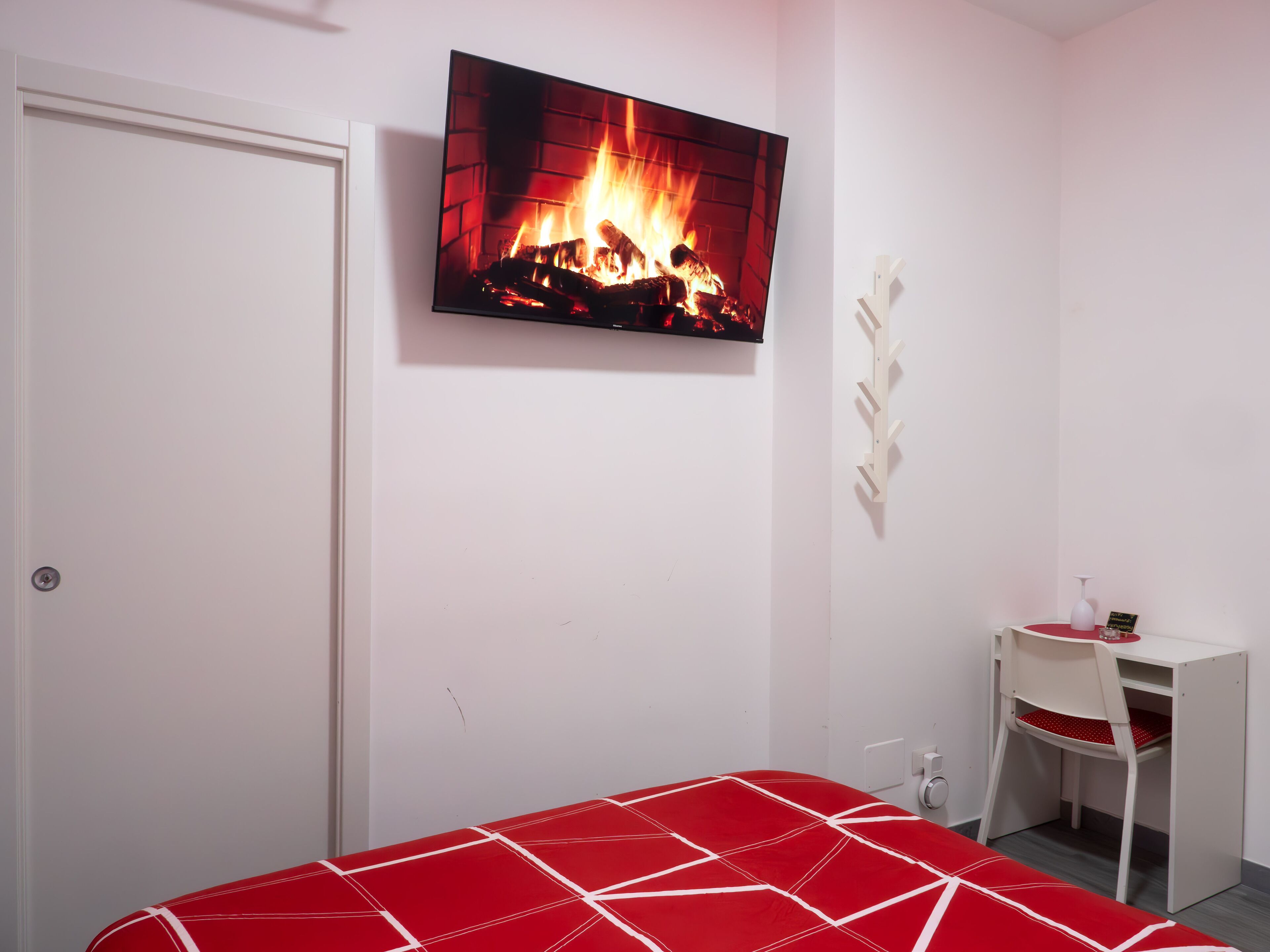 Quarto com cama, escrivaninha e TV fixada na parede exibindo uma lareira.
