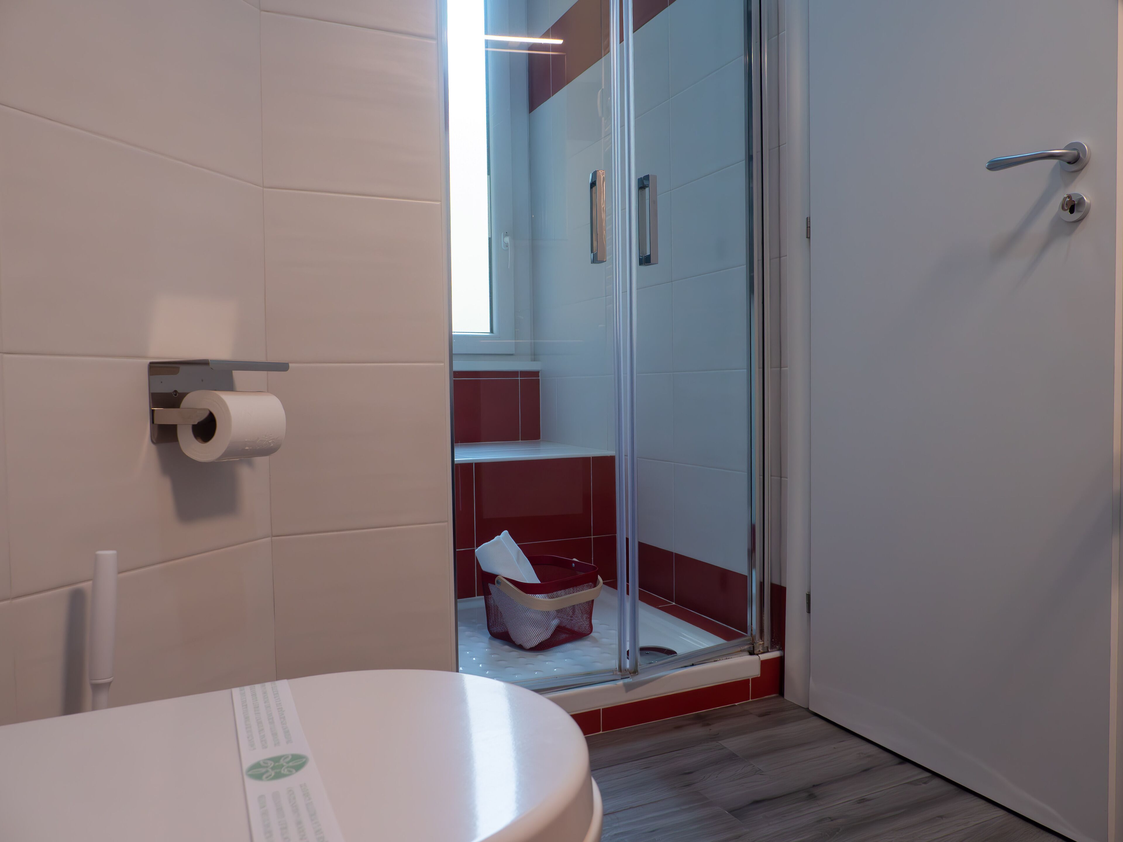 A bathroom with a toilet, a paper towel dispenser, a glass shower enclosure, and a red basket.