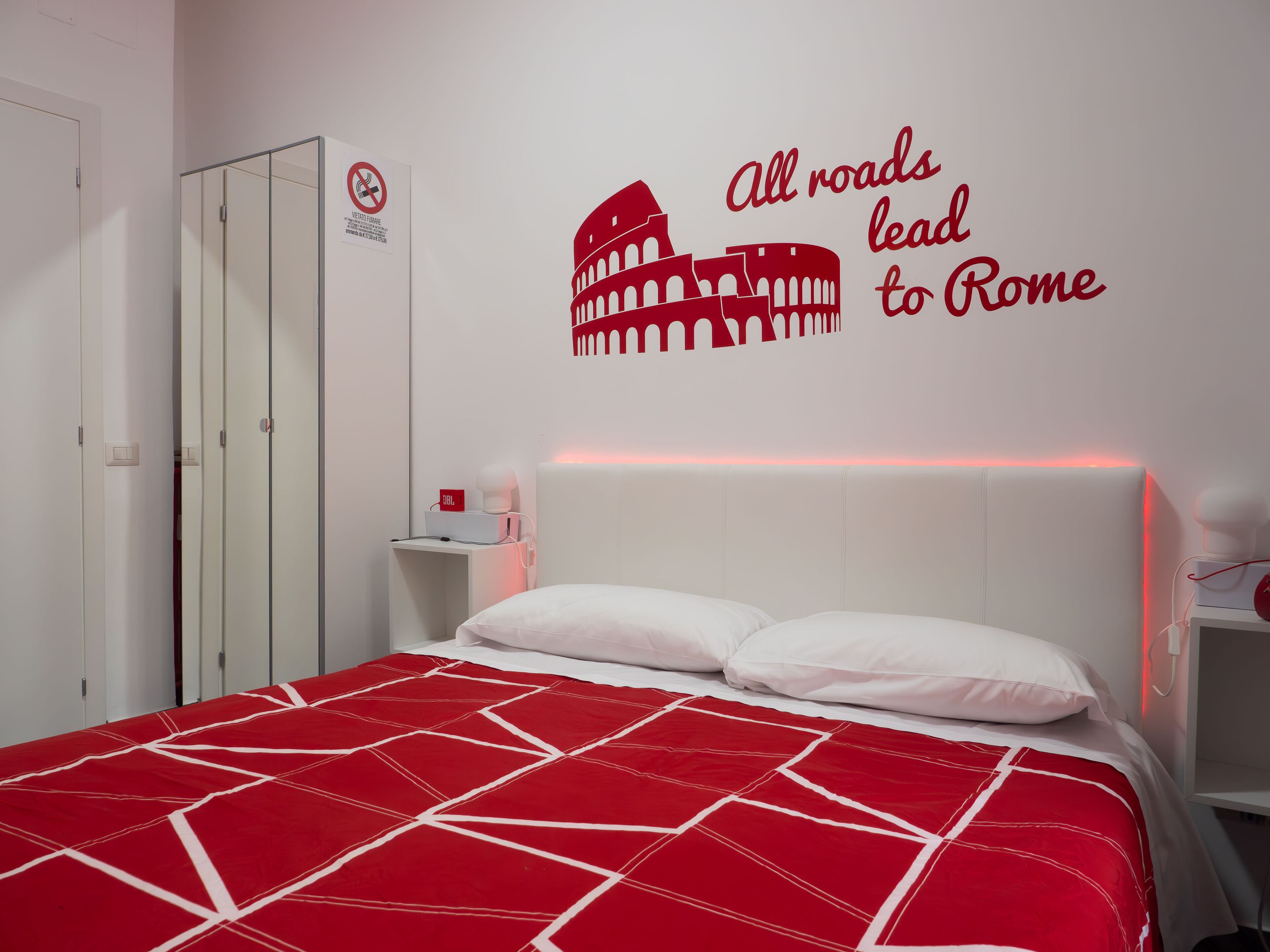 Une chambre d’hôtel avec une couvre-lit aux motifs géométriques rouge et blanc, un autocollant mural représentant le Colisée avec l’inscription « Toutes les routes mènent à Rome », une armoire blanche et une table de chevet avec une lampe et un objet rouge.