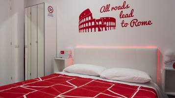 Une chambre d’hôtel avec une couvre-lit aux motifs géométriques rouge et blanc, un autocollant mural représentant le Colisée avec l’inscription « Toutes les routes mènent à Rome », une armoire blanche et une table de chevet avec une lampe et un objet rouge.