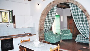 A kitchen with a dining table, chairs, and a checkered curtain over an archway leading to a living room with a patterned sofa. - Villa Albergotti (Arezzo)