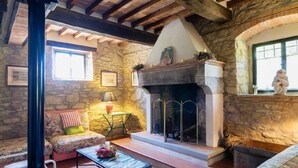 A rustic stone interior with a fireplace, wooden beams, a cushioned seating area, and a small table with a vase. - Villa Albergotti (Arezzo)