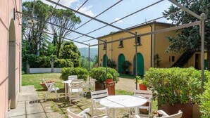 An outdoor patio with white chairs and tables, surrounded by greenery and a building with green arches. - Villa Albergotti (Arezzo)
