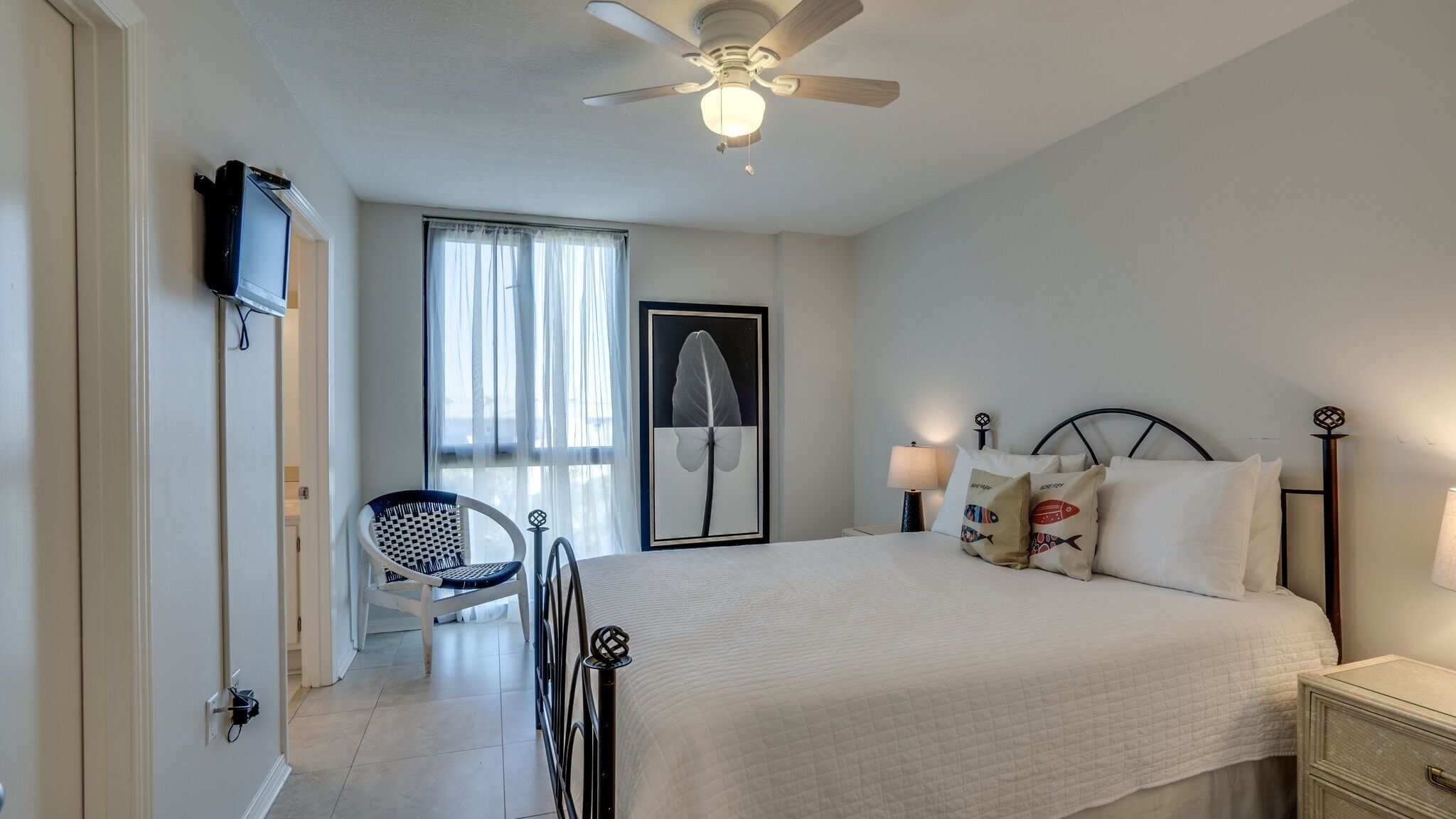 A bedroom with a bed, a chair, a ceiling fan, a TV, and a window with curtains.