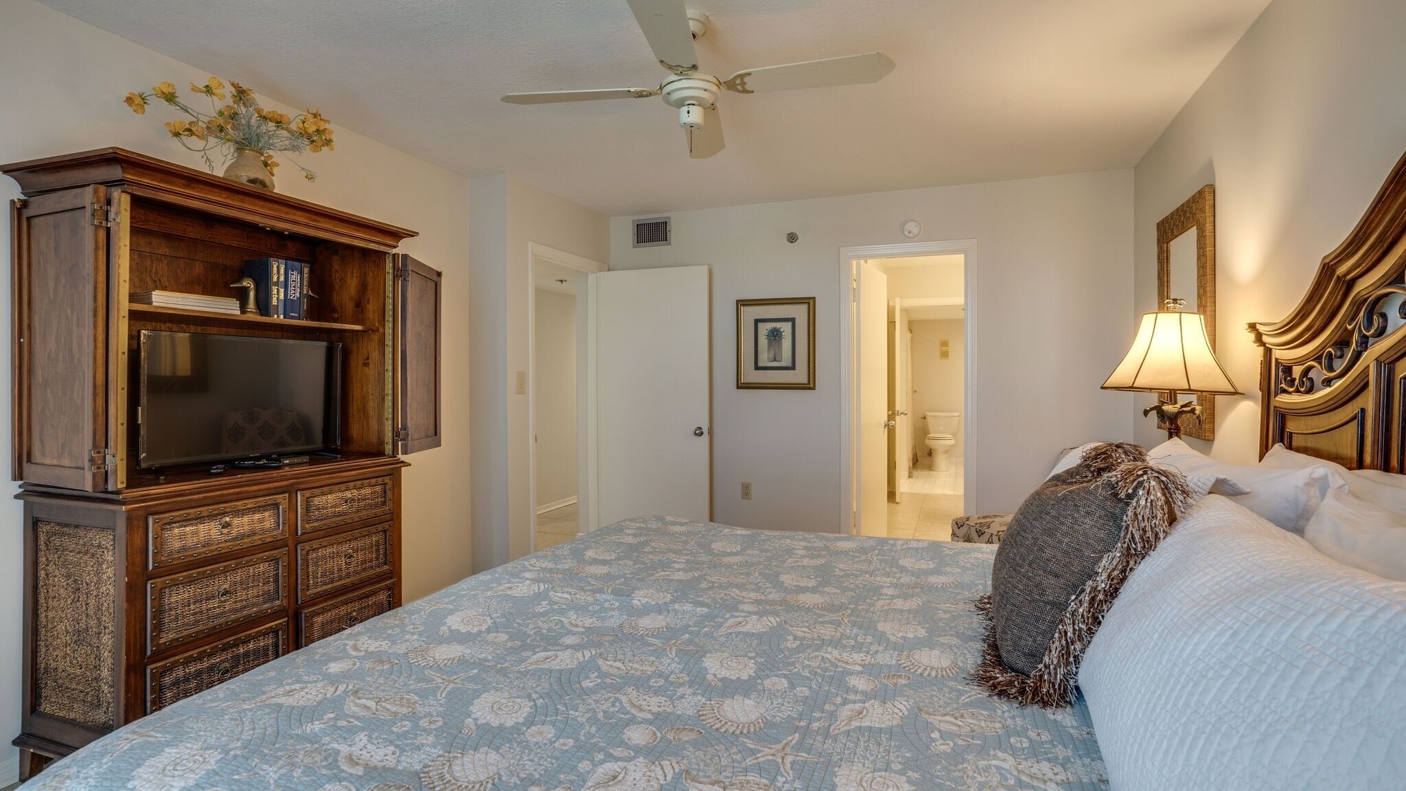 A bedroom with a large bed, a wooden cabinet with a television, a ceiling fan, and a door leading to a bathroom.