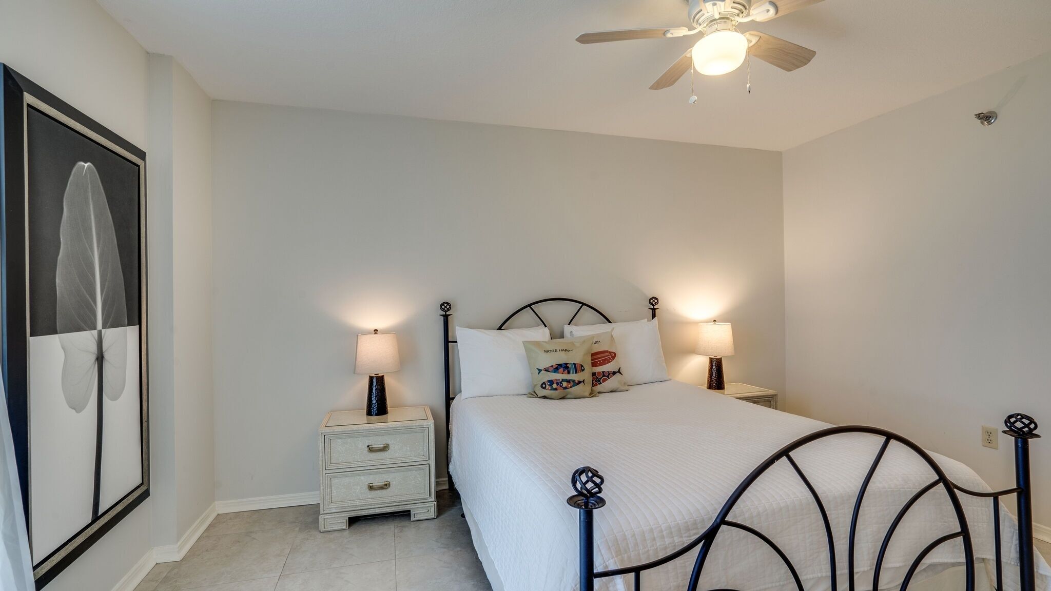 Une chambre à coucher comprenant un lit, deux tables de chevet avec des lampes, un ventilateur de plafond et une œuvre d’art encadrée accrochée au mur.